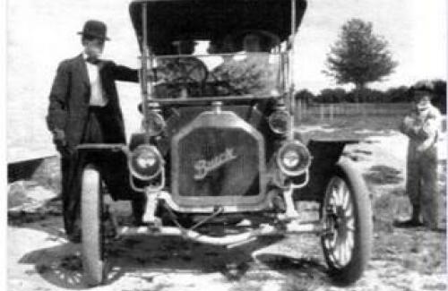 A black and white photo of a man in a bowler hat standing next to an early 1900's Buick