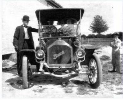 A black and white photo of a man in a bowler hat standing next to an early 1900's Buick