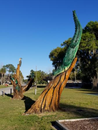 Chainsaw carving of a green alligator sliding down a honey gold tree trunk, another carving in the background